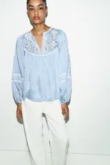 Blusa de rayas verticales blancas y celestes, con cuello redondo y escote en pico. Presenta detalles de bordados perforados y puntillas en el pecho y las mangas. Tiene mangas largas abullonadas con puños elásticos y cierre frontal con botones forrados.