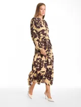 Vestido maxi de crepe con estampado floral en tonos beige y marrón oscuro. Presenta escote en V con botones frontales, mangas largas amplias con puños abotonados y cintura ajustable mediante cordón trenzado. La falda cuenta con un diseño de vuelos escalonados que aporta movimiento.