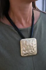 Collar de diseño con dije cuadrado de gran tamaño en tono dorado con textura irregular y martillada, suspendido de múltiples cordones negros finos.
