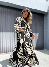 Vestido largo tipo túnica con estampado de hojas en contraste blanco y negro. Presenta cuello camisero con botones, mangas largas amplias y falda con volados escalonados.