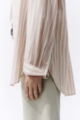 Camisa de corte oversize, con diseño de rayas verticales en tonos rosados, blancos y beiges, cuello clásico, mangas largas con puños abotonados y cierre frontal de botones.
