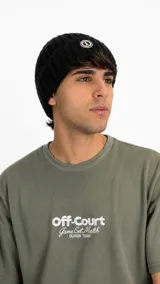 Gorro tipo beanie de punto acanalado en color negro, con diseño de doblez y parche circular bordado en la parte frontal.