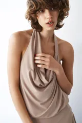 Vestido midi de color beige con cuello halter y escote drapeado. Presenta un diseño con espalda descubierta y falda con bajo asimétrico.