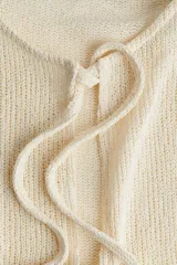 Top corto sin mangas de punto color blanco roto, con escote redondo y abertura frontal que se cierra mediante tres lazos anudados. Presenta un detalle de peplum con ligero vuelo en el bajo.