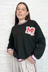 Buzo negro de rústico con cuello, puños y ruedo con ribete rosa y doble línea negra. Estampado en el pecho con la letra M en rojo y blanco, decorada con flores y un tigre.