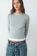 Camiseta de manga larga con diseño de doble capa, confeccionada en jersey suave y elástico. Presenta un calce ajustado, cuello redondo y un estilo minimalista que combina una capa superior en color verde grisáceo con una capa inferior en color blanco.