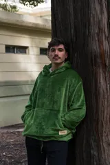 Buzo de textura peluda y suave en color verde, con capucha amplia, bolsillo frontal tipo canguro y cordón ajustable en el ruedo que permite modificar el largo de la prenda.