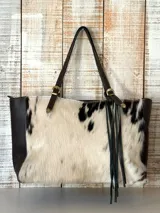 Cartera tipo tote confeccionada en cuero con frente de piel vacuna con manchas en blanco y negro. Presenta laterales y correas de cuero marrón oscuro con hebillas metálicas decorativas y flecos colgantes en uno de los laterales.