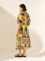 Vestido midi con estampado de piñas y motivos geométricos en tonos amarillo, marrón y negro. Presenta escote en V, mangas cortas y corte en la cintura con aberturas laterales.