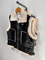 Chaleco estilo gamulán de cuero sintético negro con interior y bordes de corderito sintético color crema. Presenta cierre frontal, bolsillos laterales amplios y detalles de hebillas ajustables en los costados.