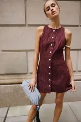 Vestido corto de denim color bordó, sin mangas, con cuello redondo, costuras pespunteadas a contratono y cierre frontal con fila de botones metálicos.