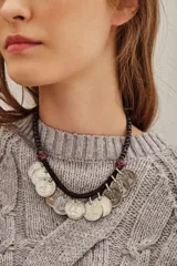 Collar corto de estilo étnico con cordón negro de poliéster y múltiples dijes colgantes en forma de monedas de metal plateado. Presenta detalles de cuentas rojas decorativas a los lados.
