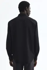 Camisa de vestir negra con cuello inglés, tapeta clásica y mangas largas con puños ajustables. Presenta un corte entallado que se ajusta al cuerpo y bajo levemente redondeado.