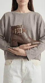 Bota corta de cuero con taco cuadrado bajo y punta redondeada. Presenta una tira decorativa en el tobillo con una hebilla metálica rectangular. Cuenta con cierre lateral interno para facilitar el calce.