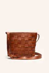 Cartera tipo morral color tostado, confeccionada en cuero con diseño trenzado artesanal, forma de balde y tira regulable para llevar al hombro o cruzada.