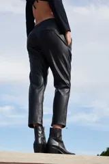 Pantalón sastrero de ecocuero negro, con corte recto, bolsillos delanteros y traseros, y botamanga irregular más corta en la parte frontal.