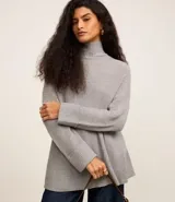 Suéter de punto alargado color gris, con cuello alto ajustado, mangas largas ajustadas y detalle abierto traspasado en la espalda.