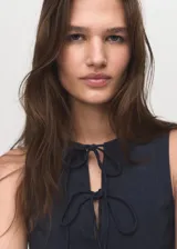 Vestido corto sin mangas con efecto denim, cuello redondo y detalle de lazos frontales. Presenta un diseño con tablas y cierre de cremallera en la parte posterior.