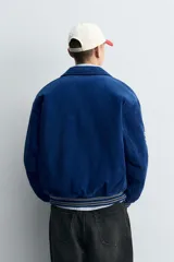 Cazadora estilo varsity confeccionada en pana de algodón azul, con interior ligeramente acolchado. Presenta cuello solapa, cierre frontal con botones a presión, bolsillos laterales de vivo y acabados en rib a rayas en puños y cintura. Incluye múltiples parches bordados decorativos en el frente.