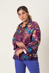 Blusa tipo casaca italiana abotonada, de satén estampado con fondo violeta y flores en colores azul, mostaza y naranja. Tiene cuello de camisa y mangas largas con presilla para sujetar.