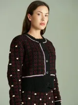 Cardigan de punto negro y rojo con diseño texturizado, cuello redondo y cierre frontal con botones. Presenta detalles de ribetes en blanco y violeta en el cuello, la botontera, los puños y el ruedo, que cuenta con flecos colgantes.
