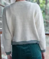 Sweater artesanal de lana merino en color natural, con diseño tejido de jacquard en la parte inferior y bordado a mano de dos cisnes enfrentados formando un corazón en el pecho. Presenta cuello redondo, puños y ruedo en tono verde contrastante.