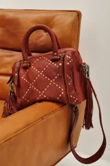 Bolso de mano tipo doctor bag o bowling bag, color marrón cuero, con diseño acolchado en rombos y tachas doradas. Posee doble asa de mano y un dije colgante de flecos largos de cuero marrón.