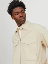 Chaqueta color crema con cuello camisero, cierre frontal con cremallera metálica, bolsillos de parche con solapa abotonada en el pecho y corte recto.