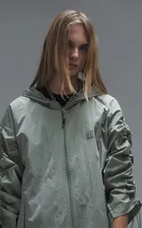 Campera rompevientos de diseño oversize, color verde, confeccionada en microfibra técnica ligera. Presenta capucha integrada, cierre frontal técnico y un sistema de frunces elásticos regulables a lo largo de las mangas que permiten ajustar el volumen de la prenda.