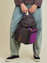 Mochila marrón con detalles en violeta. Tiene un bolsillo frontal superior con cierre, un bolsillo lateral de red y el logo de Rusty Worldwide estampado en naranja.