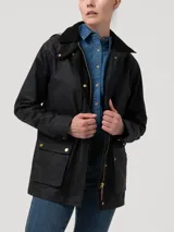Chaqueta de algodón orgánico encerado, resistente al agua y al viento, con diseño de corte recto. Presenta cuello alto, gorro removible con forro a cuadros, cierre frontal metálico oculto por solapa con botones a presión y dos bolsillos frontales con solapa. Incluye detalles en cotelé y forro interior funcional.