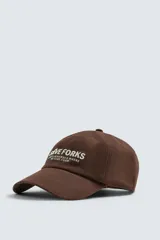 Gorra de seis paneles confeccionada en tejido de algodón, color marrón, con bordado frontal en color crema que incluye texto. Cuenta con ajuste en la parte posterior.