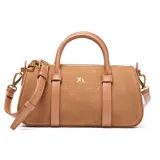 Cartera tipo baúl compacta de símil gamuza color tostado, combinada con detalles en PU liso del mismo tono. Cuenta con doble asa corta rígida, correa larga regulable y desmontable, y herrajes metálicos dorados. El compartimento principal tiene cierre superior y un bolsillo interno con cierre. Presenta el logo XL metalizado en la parte frontal.