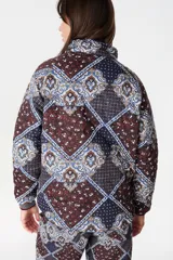 Campera de abrigo con cuello alto y manga ranglan, confeccionada en tela capitoneada con relleno y forro interno. Presenta un diseño estampado con motivos geométricos y paisley en tonos azules y marrones.