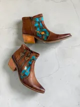 Botas cortas de cuero marrón terracota con diseño pintado a mano en la parte frontal en tonos turquesa y dorado. Cuentan con taco cuadrado de madera, doble cierre lateral con tiradores metálicos personalizados y punta fina.
