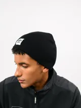 Gorro tipo beanie de tejido de punto acanalado en color verde oliva, con parche frontal rectangular blanco que presenta el logo de Umbro en rojo.