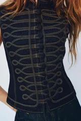 Chaleco de denim azul oscuro con cuello redondo y cierre frontal de botones. Presenta un diseño de pespuntes decorativos en contraste con forma de alamares en la parte delantera.
