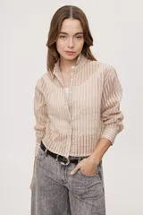 Camisa de manga larga confeccionada en tejido semitransparente con diseño de rayas verticales en tonos beige, azul y blanco. Presenta cuello clásico y cierre frontal con botones.