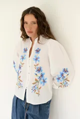 Camisa blanca de manga larga abullonada, con delicados bordados florales en tonos azules y verdes a lo largo de la botonadura frontal y en las mangas. Presenta cuello mao con reborde en contraste y cierre frontal con botones.