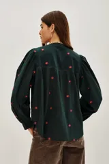 Camisa de algodón color crudo con bordados florales pequeños en azul marino. Presenta mangas largas abullonadas, cuello redondo y una abertura frontal con botones y detalles de encaje calado en la pechera y hombros.