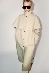 Trench de cuello con solapa y manga larga, con detalle de capa desmontable del mismo tejido en la parte superior. Cuenta con cierre frontal de botones y bolsillos en el delantero.
