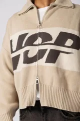 Campera tejida de punto con cierre frontal completo y cuello polo. Presenta un diseño de franja horizontal ancha en color crema con estampado gráfico oscuro sobre una base de color marrón. Posee puños y ruedo acanalados, con un pequeño parche de marca en el puño izquierdo.