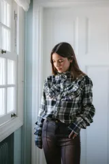 Camisa de franela a cuadros en tonos negro, blanco y gris, con cuello alto y detalle de volados en el frente y puños. Presenta cierre frontal con botones.