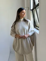 Poncho de tejido de punto color beige con cuello alto y diseño de trenzas verticales en el frente. Presenta mangas integradas con puños acanalados y un corte holgado con ruedo redondeado.
