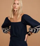 Blusa corta azul marino con escote recto estilo hombro a hombro, mangas largas abullonadas con detalle de bordado decorativo en cuerda clara.
