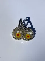 Aros colgantes con forma circular, con strass plateado alrededor y flor de vidrio amarilla con centro rojo en el interior. El arito es de acero quirúrgico.