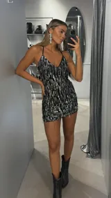 Vestido corto de fiesta con escote en V y tirantes finos. Presenta una base negra cubierta con flecos de lentejuelas plateadas que crean un efecto brillante y dinámico.