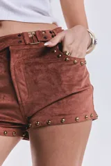 Short corto de tiro bajo confeccionado en gamuza elastizada color marrón camel. Presenta detalles de tachas metálicas doradas en los bolsillos delanteros y en los bordes inferiores, además de una faja en la cintura con hebillas metálicas laterales.