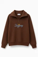 Buzo de cuello alto con cierre frontal, confeccionado en tela de jogging con felpa interior. Presenta un bordado central con la palabra Uruguay en color celeste y un sol amarillo. Posee puños y ruedo acanalados.