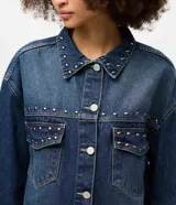 Camisa de jean estilo overshirt con corte oversized, cuello clásico y mangas largas. Presenta cierre frontal con botones, dos bolsillos delanteros con solapa y aplicación de tachuelas metálicas en el cuello y la parte superior del pecho.